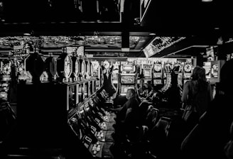 A dimly lit casino filled with rows of slot machines, some occupied by people. The machines' lights contrast with the dark surroundings, creating a vibrant yet subdued atmosphere.