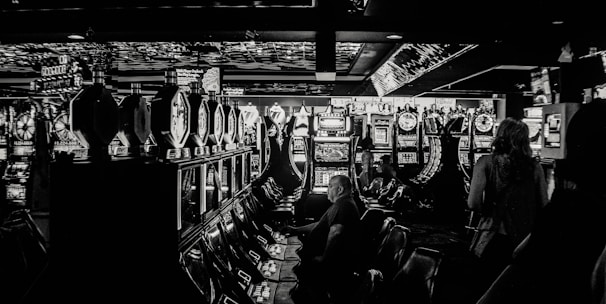 A dimly lit casino filled with rows of slot machines, some occupied by people. The machines' lights contrast with the dark surroundings, creating a vibrant yet subdued atmosphere.