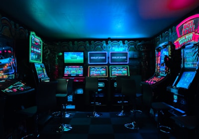 Dimly lit gaming arcade featuring a variety of brightly illuminated slot machines and video gaming screens. Neon signs and colorful displays create a vibrant atmosphere. The arrangement of gaming machines suggests an inviting space for entertainment, with chairs positioned for players.