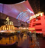 A bustling nighttime scene with illuminated casino signage towering over a busy pedestrian street. Neon lights in red and yellow highlight the names of various establishments. People are walking in different directions, enjoying the vibrant atmosphere.