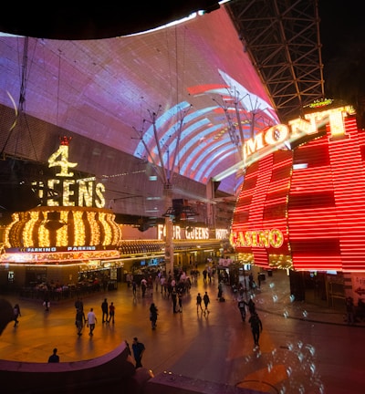 A bustling nighttime scene with illuminated casino signage towering over a busy pedestrian street. Neon lights in red and yellow highlight the names of various establishments. People are walking in different directions, enjoying the vibrant atmosphere.