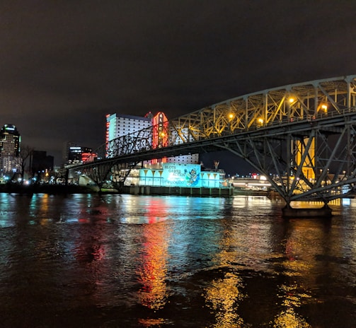 A brightly lit casino with neon signs and red lights is reflected in a river. A bridge stretches across the water, with an urban skyline visible in the background against an overcast night sky.