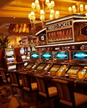 A casino floor featuring rows of brightly lit video poker machines. The area is illuminated by elegant chandeliers, with a display board showing a large sum of money, suggesting a possible jackpot. The rich decor includes luxurious carpets and comfortable seating, adding to the opulent atmosphere.