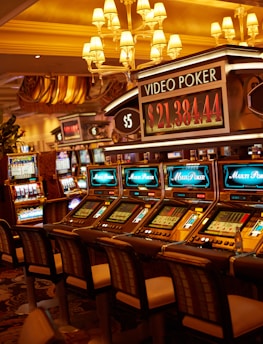 A casino floor featuring rows of brightly lit video poker machines. The area is illuminated by elegant chandeliers, with a display board showing a large sum of money, suggesting a possible jackpot. The rich decor includes luxurious carpets and comfortable seating, adding to the opulent atmosphere.