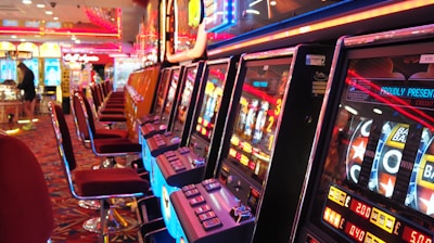 A brightly lit casino interior featuring a row of colorful slot machines. The neon lights create a vibrant atmosphere, with patterned carpeting adding to the decor. Stools are aligned neatly in front of each machine.