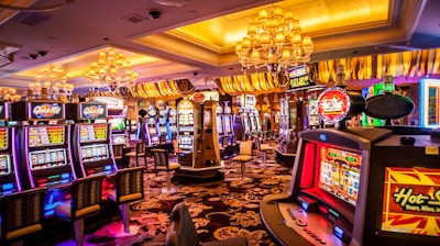 A vibrant casino interior filled with rows of colorful slot machines, each illuminated with bright lights and displays. The ceiling is adorned with elegant chandeliers and golden drapes, creating a luxurious atmosphere. The patterned carpet adds to the elaborate design of the space.