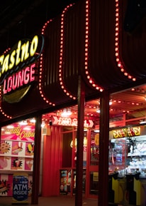 A brightly lit casino lounge entrance adorned with neon lights. The exterior features colorful signage including the words 'Casino' and 'Lounge' in vivid colors, with additional lights framing the building. Inside, a glimpse of a claw machine filled with plush toys and arcade games is visible. The area exudes an inviting yet bustling atmosphere typical of entertainment venues.