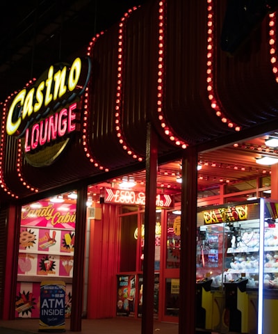 A brightly lit casino lounge entrance adorned with neon lights. The exterior features colorful signage including the words 'Casino' and 'Lounge' in vivid colors, with additional lights framing the building. Inside, a glimpse of a claw machine filled with plush toys and arcade games is visible. The area exudes an inviting yet bustling atmosphere typical of entertainment venues.