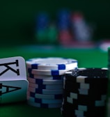 A casino scene featuring two playing cards, the ace and king of clubs, partially concealed by a player's hand. Nearby, there are stacks of multicolored poker chips on a green felt table, with blurred colorful background elements suggesting other chip stacks or cards.