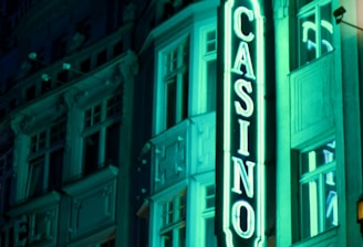 A vibrant neon sign displaying the word 'CASINO' is prominently illuminated against the facade of a building, showcasing intricate architectural details and a classic design. The sign emits a bright teal glow, casting light on the surrounding area and creating a lively atmosphere at night.
