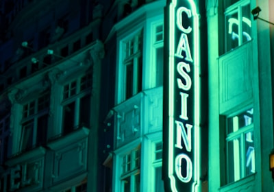 A vibrant neon sign displaying the word 'CASINO' is prominently illuminated against the facade of a building, showcasing intricate architectural details and a classic design. The sign emits a bright teal glow, casting light on the surrounding area and creating a lively atmosphere at night.