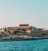 A seaside view of a large building labeled as a casino, situated on a rocky shore. The building is surrounded by palm trees and other smaller structures. The calm sea water reflects the sky, and there are several closed umbrellas lined along the waterfront.