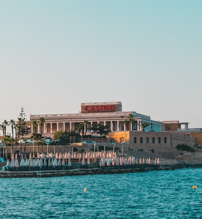 A seaside view of a large building labeled as a casino, situated on a rocky shore. The building is surrounded by palm trees and other smaller structures. The calm sea water reflects the sky, and there are several closed umbrellas lined along the waterfront.