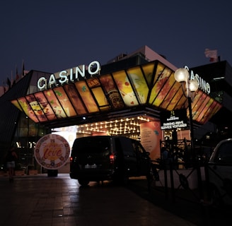 A building with a brightly illuminated entrance is seen at night. The sign above reads 'Casino' in bright letters. The facade features colorful graphics depicting various indulgent themes. Several vehicles are parked near the entrance, and people are visible in the vicinity. Tall lamp posts with spherical lights are present, casting a warm glow.