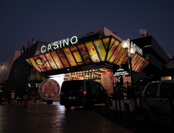 A building with a brightly illuminated entrance is seen at night. The sign above reads 'Casino' in bright letters. The facade features colorful graphics depicting various indulgent themes. Several vehicles are parked near the entrance, and people are visible in the vicinity. Tall lamp posts with spherical lights are present, casting a warm glow.
