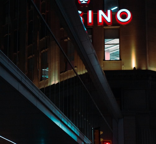 A night scene featuring a building with illuminated neon signage displaying 'CASINO'. The environment is urban with reflections on glass windows and glowing street traffic lights below. The atmosphere is dimly lit, with artificial lighting casting vibrant colors.