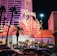 A brightly lit casino with neon lights in vibrant red, pink, and blue, featuring a large flamingo signage. Palm trees are visible in the foreground with vehicles passing by on the street at night.