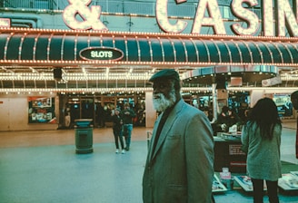 An older man with a gray beard and wearing a suit and hat stands in front of a brightly lit casino. The background features a large illuminated sign reading 'CASINO' and 'SLOTS' among other words, with numerous lights outlining the entrance. Several other people are visible in the background, engaged in various activities, some walking, some standing, and one looking at something on display.