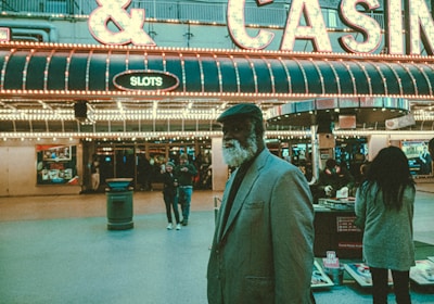 An older man with a gray beard and wearing a suit and hat stands in front of a brightly lit casino. The background features a large illuminated sign reading 'CASINO' and 'SLOTS' among other words, with numerous lights outlining the entrance. Several other people are visible in the background, engaged in various activities, some walking, some standing, and one looking at something on display.