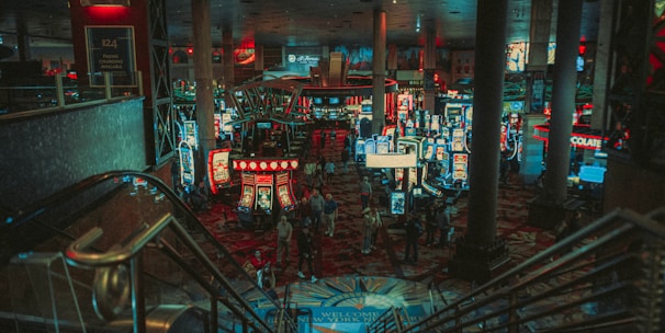 A bustling casino floor filled with groups of slot machines and people walking around. The atmosphere is vibrant with neon lights and illuminated signs. The interior has a ceiling supported by large pillars and there are stairs leading down to the gaming area.
