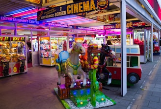 A vibrant amusement arcade featuring various games and bright neon signs. Mechanical horse rides are in the foreground, surrounded by claw machines with plush toys. The entrance is adorned with signs for a cinema and arcade, creating a lively and colorful atmosphere.