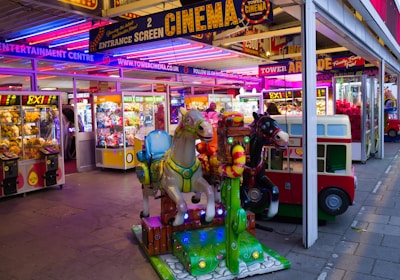 A vibrant amusement arcade featuring various games and bright neon signs. Mechanical horse rides are in the foreground, surrounded by claw machines with plush toys. The entrance is adorned with signs for a cinema and arcade, creating a lively and colorful atmosphere.