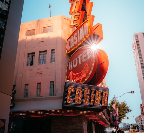 A vibrant city scene featuring a tall, illuminated casino sign with retro-style neon lights. The sign dominates the building facade with the words 'CASINO' and 'HOTEL'. Sunlight peeks through, creating a dramatic lens flare effect. The street is lively, with pedestrians casually walking and a hint of urban hustle.